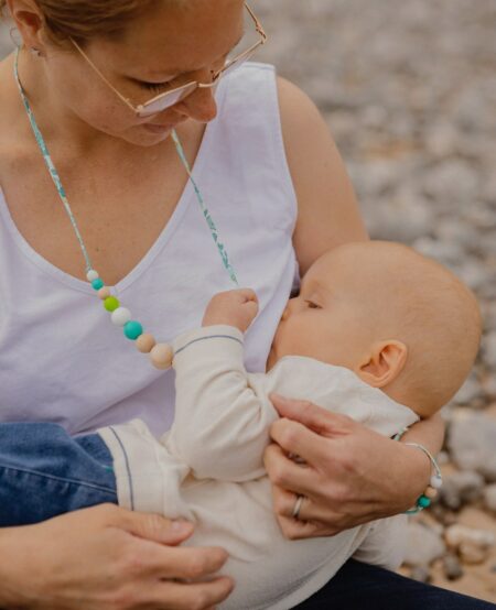 Bébé qui tète sa maman en tenant le collier d'allaitement bora bora avec des perles bleus blanches et bois